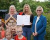 Zu sehen sind drei blondhaarige Frauen. Die Frau in der Mitte hält eine Urkunde in der Hand. Davor stehen mehrere Kinder im Vorschulalter.