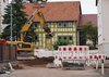 Zu sehen ist eine Straß mit Häusern im Hintergrund. Ein Bereich der Straße ist abgesperrt. Dahinter ist ein gelber Bagger.