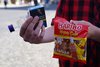 Zu sehen ist eine Nahaufnahme von den Händen einer Person. Sie hält eine Tüte Gummibärchen, Alkohol und eine Schachtel Zigaretten.