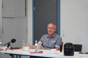 Zu sehen ein Mann. Er sitzt an einem Tisch und spricht. 