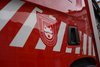 Zu sehen ist ein Teil reines roten Feuerwehr-Einsatzwagens. Darauf ist eine Folierung mit dem einem weißen Wappen bzw. Logo. 