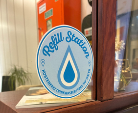 Zu sehen ist ein blauer Aufkleber auf einer Glasscheibe. Darauf ist ein blauer Wassertropfen und die Worte "Refill-Station. Kostenfrei Trinkwasser. Free tap water" zu sehen.