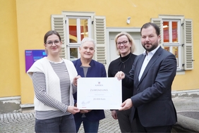 Drei Frauen und ein Mann stehen vor einer gelben Hauswand zusammen. Sie halten einen symbolischen Scheck in der Hand und lachen in die Kamera.