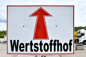 Ein Schild mit einem roten Pfeil drauf. Darunter steht in schwarzer Schrift "Wertstoffhof".