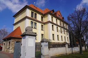 Zu sehen ist eine massives Gebäude mit gelber Wandfarbe. Davor ist ein ein Zaun aus Stein.