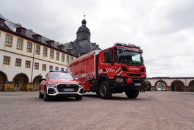 Dies ist eine weite Außenaufnahme, die einen roten Feuerwehrwagen und einen roten Audi SUV zeigt, beide mit Blaulicht ausgestattet, geparkt auf einer gepflasterten Fläche vor einem beeindruckenden, historischen Gebäudekomplex. Der Himmel ist bewölkt.
