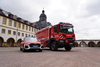 Dies ist eine weite Außenaufnahme, die einen roten Feuerwehrwagen und einen roten Audi SUV zeigt, beide mit Blaulicht ausgestattet, geparkt auf einer gepflasterten Fläche vor einem beeindruckenden, historischen Gebäudekomplex. Der Himmel ist bewölkt.