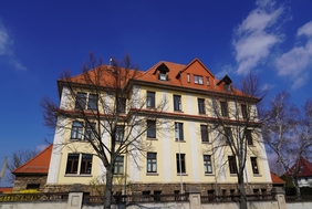 Zu sehen ist ein mehrstöckiges, gelbes Haus. Davor stehen zwei Bäume ohne Blätter. 
