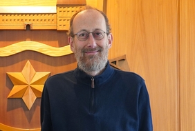 Vor einem hölzernen Hintergrund steht ein Mann mit Brille und dunklem Oberteil. Er lächelt in die Kamera.