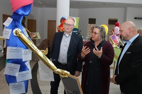 Drei Personen stehen vor einer bunten Puppe und unterhalten sich. An der Puppe heften bunte Zettel mit Informationen, Fragen und Erfahrungen zum Thema Demenz.