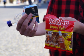 Zu sehen ist eine Nahaufnahme von den Händen einer Person. Sie hält eine Tüte Gummibärchen, Alkohol und eine Schachtel Zigaretten.