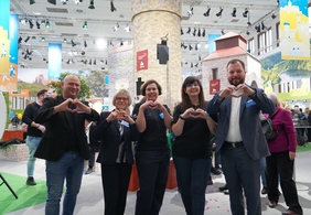 Zu sehen sind fünf Menschen, die nebeneinander stehen und in die Kamera lächeln. Alle formen mit den Händen ein Herz.