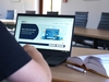 Zu sehen ist eine Person von hinten, die an einem Laptop sitzt. Auf dem Bildschirm ist die Cloud der VHS geöffnet. Neben dem Laptop auf dem Tisch liegt ein offenes Buch.
