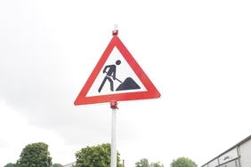 Ein Verkehrsschild mit einem Bauarbeiter steht an einer Straße. Im Hintergrund ist ein grauer Himmel zu sehen.