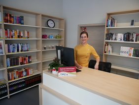 Zu sehen ist eine Frau an einem Tresen. Hinter ihr stehen Regale mit Büchern.