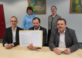 Zu sehen sind fünf Personen, zwei Frauen und drei Männer, die in die Kammer schauen. Einer der Männer hält einen unterschriebenen Vertrag in die Kamera. 