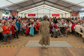 Zu sehen ist eine Sängerin von hinten, wie sie vor einem Publikum auf der Bühne steht und singt. Das Publikum besteht vor allem auf Seniorinnen und Senioren. Die Veranstaltung findet in einem Festzelt statt.