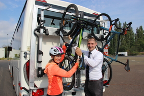 Zu sehen sind eine Frau mit Helm und ein Busfahrer. Gemeinsam befestigen sie das Rad am Busheck und schauen dabei in die Kamera.