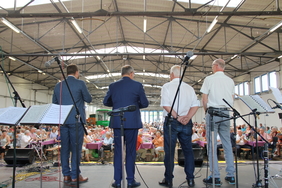 Vier Männer stehen auf einer Bühne in der Wagenhalle. Sie sind von hinten zu sehen. Vor ihnen sind Tische aufgebaut, auf denen Seniorinnen und Senioren sitzen.