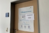 Zu sehen ist eine verschlossene Tür. An der Tür hängt ein Plakat, dass den Passbildautomaten erklärt, darüber ist ein weiteres Schild geklebt. Darauf steht, dass der Automat defekt ist. 