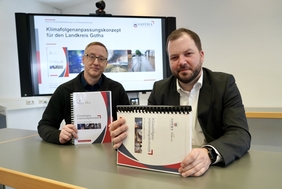 Zu sehen sind zwei Männer, die in die Kamera schauen. Beide sitzen an einem Tisch und halten ein dickes Buch mit Ringbindung in der Hand: Das Klimafolgenanpassungskonzept.