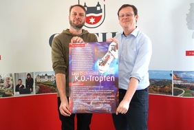 Zwei Männer stehen nebeneinander. Sie halten zusammen ein Plakat in den Händen. Auf dem Plakat ist zu lesen: "Lass dich nicht K.O.-Tropfen".