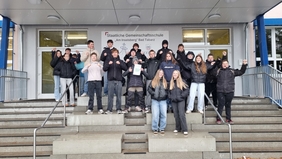 Zu sehen ist eine Gruppe Jugendliche vor der Eingangstür einer Schule. Die Jugendlichen freuen sich.