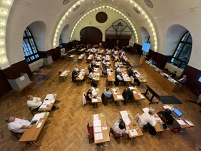 Zu sehen ist die Aula der Arnoldischule. Auf dem Parkettboden stehen angeordnet nach Fraktionen Tische. Jeweils zwei Kreistagsmitglieder sitzen an einem Tisch. 