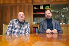 Zwei Männer sitzen nebeneinander an einem Tisch und schauen in die Kamera.