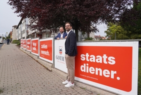 Zu sehen ist ein Zaun an dem große, rote Schilder mit den Worten "Staatsdiener", "Zahlenmensch" und "Alltagsheldin" zu lesen sind. Ein Mann steht vor dem Zaun, ein zweiter dahinter. Beide lächeln in die Kamera.