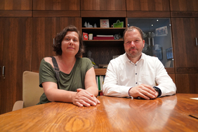 Zu sehen sind ein Mann und eine Frau, die nebeneinander an einem Holztisch sitzen und in die Kamera lächeln. 