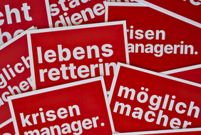 Zu sehen sind viele rote Postkarten auf denen Begriffe wie "Lebensretterin", "Möglichmacher" und "Krisenmanager" stehen.