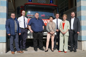 Sechs Personen stehen nebeneinander und schauen in die Kamera. Im Hintergrund ist ein rotes Einsatzfahrzeug der Feuerwehr in einer Halle zu sehen.
