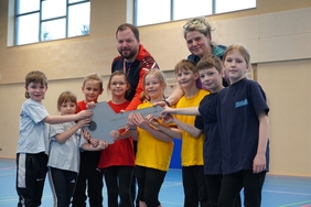 Mehrere Kinder stehen in einer Reihe in einer Sporthalle. Hinter ihnen stehen ein Mann und eine Frau. Gemeinsam halten sie alle einen großen Schlüssel in der Hand.