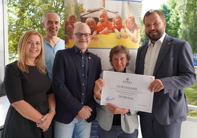 Fünf Menschen stehen nebeneinander. Eine Frau hält zusammen mit einem Mann eine Urkunde in der Hand auf der unter anderem 126.000 Euro steht. Alle lächeln in die Kamera.