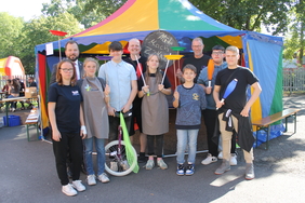 Zu sehen ist ein kleines Zirkuszelt. Davor stehen Kinder und Erwachsene mit Utensilien aus dem Zirkus. Dazu gehören ein Einrad, Jonglierkeulen und vieles mehr. 