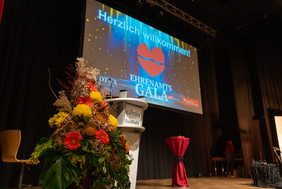 Zu sehen ist eine Bühne mit Blumen und einem Rednerpult. Im Hintergrund ist eine Präsentation, auf der steht "Herzlich Willkommen zur Ehrenamtsgala 2025".