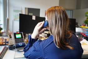 Zu sehen ist eine junge Frau, die an einem Schreibtisch sitzt und telefoniert. Sie ist nur von hinten zu sehen.