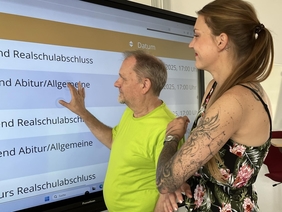 Ein Mann und eine Frau stehen vor einem Whiteboard. Auf dem Bildschirm sieht man Informationen zu den Kursen der KVHS.