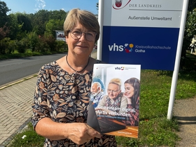 Heike Strumpf, Leiterin der Kreisvolkshochschule, hält das neue Programmheft für das Herbstsemester in den Händen. Sie steht draußen vor einem Schild, auf dem "Kreisvolkshochschule Gotha" steht.