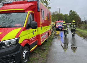Zwei Feuerwehrmänner laufen auf einer nassen, schmalen Straße. Links neben ihnen ist ein rotes Einsatzfahrzeug. Der Himmel ist komplett bewölkt und grau.