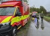 Zwei Feuerwehrmänner laufen auf einer nassen, schmalen Straße. Links neben ihnen ist ein rotes Einsatzfahrzeug. Der Himmel ist komplett bewölkt und grau.