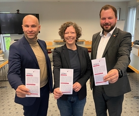 Zu sehen sind Andreas Kettritz, Simone Rieth und Landrat Onno Eckert, die nebeneinander in einem Seminarraum stehen und das Leitbild als Flyer in die Kamera halten.