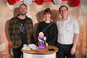 Zu sehen sind zwei Männer und eine Frau die nebeneinander stehen und in die Kamera lächeln. Vor ihnen steht ein kleiner Tisch mit Infomaterialien zu der Kampagne.