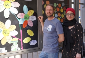 Zu sehen sind ein Mann und eine Frau vor einem Schaukasten mit bunten Zetteln. Die Zettel sind in Form einer Blume angeordnet. Darauf steht, was Jugendliche im Leben feiern.