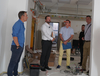Zu sehen sind vier Männer auf einer Baustelle. Die Wände sind frisch gestrichen, der Boden ist abgedeckt, es hängen Kabel von der Decke und im Hintergrund stehen Farbeimer und Werkzeuge auf dem Boden. Die vier Männer schauen sich im Raum um. 