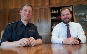 Zu sehen sind zwei Männer die gemeinsam an einem Tisch sitzen. Einer der Männer trägt eine Feuerwehr-Uniform und der andere ein weißes Hemd und weinrote Krawatte.
