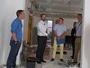 Zu sehen sind vier Männer auf einer Baustelle. Die Wände sind frisch gestrichen, der Boden ist abgedeckt, es hängen Kabel von der Decke und im Hintergrund stehen Farbeimer und Werkzeuge auf dem Boden. Die vier Männer schauen sich im Raum um. 