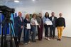 Mehrere Menschen stehen nebeneinander und halten Urkunden in den Händen. Rechts im Bild ist eine Videokamera zu sehen, die auf sie gerichtet ist.