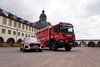 Dies ist eine weite Außenaufnahme, die einen roten Feuerwehrwagen und einen roten Audi SUV zeigt, beide mit Blaulicht ausgestattet, geparkt auf einer gepflasterten Fläche vor einem beeindruckenden, historischen Gebäudekomplex. Der Himmel ist bewölkt.